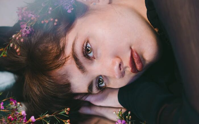 Tender young female looking away among delicate purple flowers and reflecting in mirror in soft focus skincare